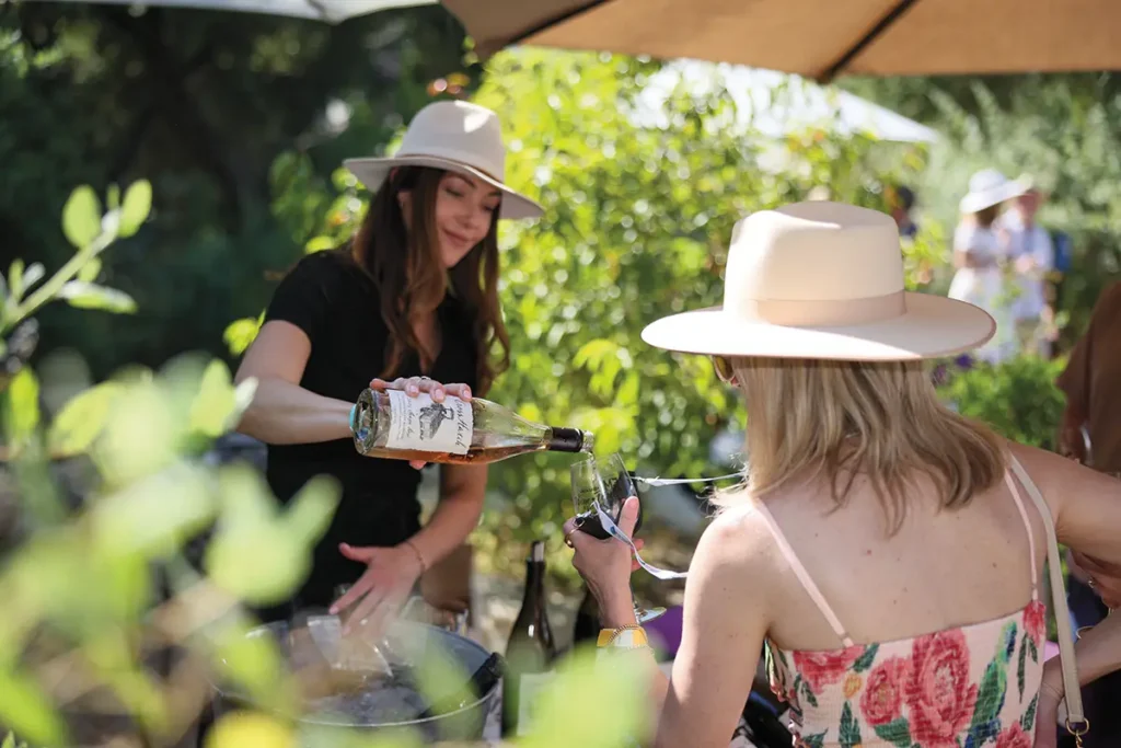 Woman in a white hat pouring wine for a guest at an outdoor wine tasting in a sunny garden setting.