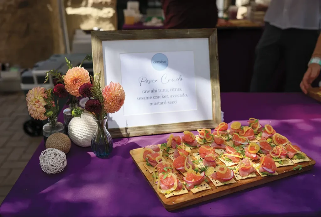 Platter of pesca crudo bites on sesame crackers with citrus, avocado and mustard seed, displayed on a purple tablecloth with a menu sign and flowers.