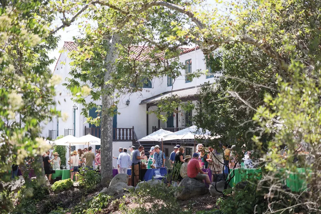 Guests gather at an outdoor event in a shaded garden courtyard with colorful tablecloths, umbrellas and a Spanish-style building in the background.
